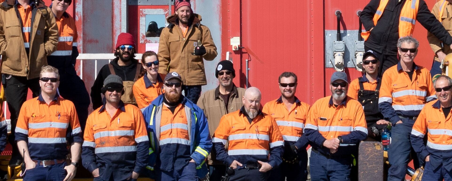 A group of expeditioners standing in front of the red workshop building all dressed in High vis work clothing.