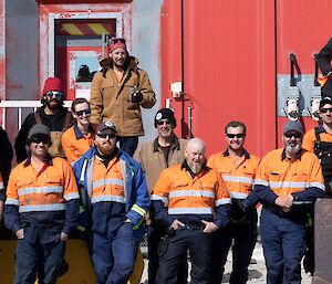 A group of expeditioners standing in front of the red workshop building all dressed in High vis work clothing.