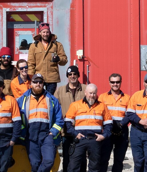 A group of expeditioners standing in front of the red workshop building all dressed in High vis work clothing.