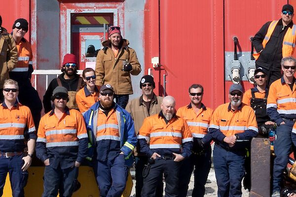 A group of expeditioners standing in front of the red workshop building all dressed in High vis work clothing.