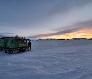 A man stands beside a Hagglund vehicle on the sea ice with a yellow sky in the distance.