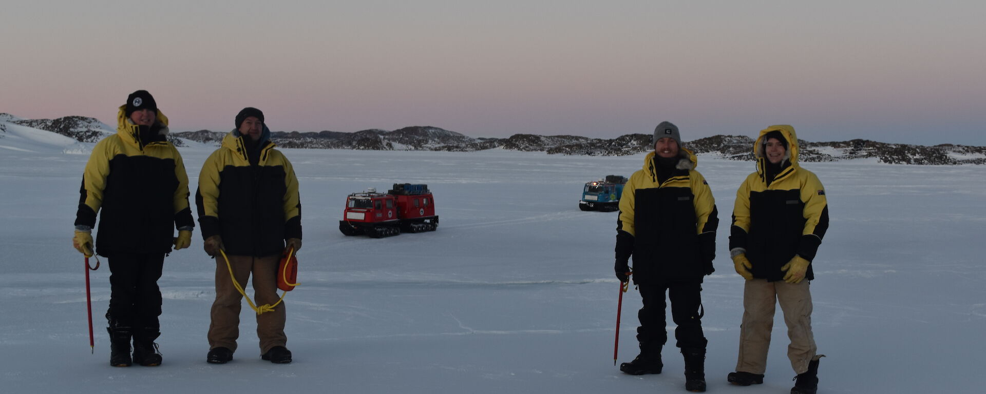 Four expeditioners in cold weather gear standing on the sea ice with a pink sunrise behind them