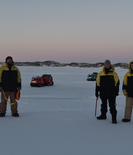 Four expeditioners in cold weather gear standing on the sea ice with a pink sunrise behind them