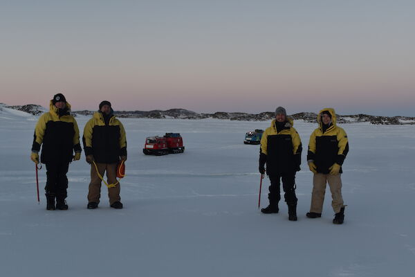 Four expeditioners in cold weather gear standing on the sea ice with a pink sunrise behind them