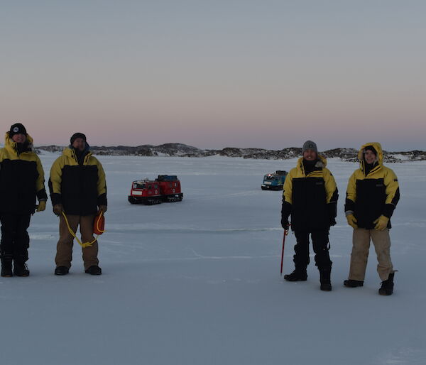 Four expeditioners in cold weather gear standing on the sea ice with a pink sunrise behind them
