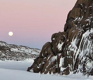 A snowy landscape image with snow dusted rocks.  A low sun in the sky is complimented by a pink sky.