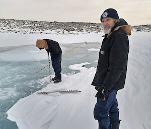 A man in cold weather gear looks to camera as a colleague nearby drills a hole in the sea ice on which they are standing.