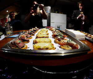 A close up image of a platter of h'or d'heurves.  Two people can be seen in the distance taking pictures of the platter.