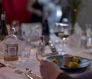 A diners place at the table with a plate scallop wantons, a white table cloth, silver service settings and a bottle of gin