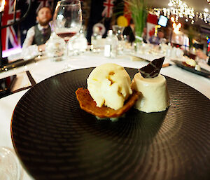 A panna cotta dessert beautifully presented on a black plate, with ice-cream and tuile