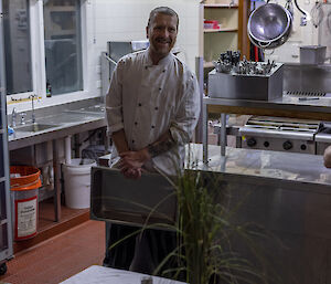 A chef in whites standing behind a decorated table set in front of a commercial kitchen, smiling to camera