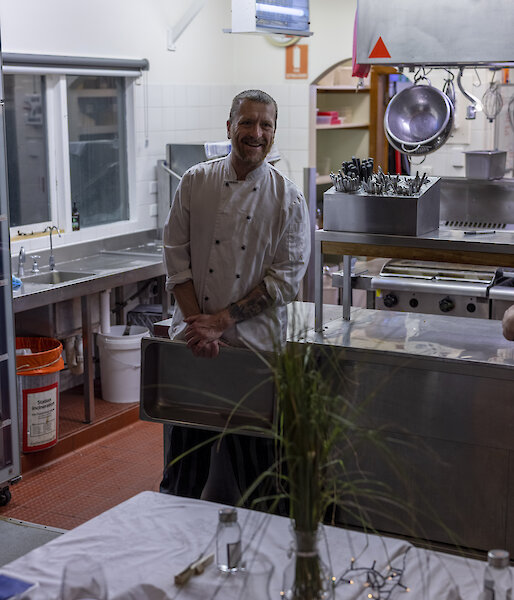 A chef in whites standing behind a decorated table set in front of a commercial kitchen, smiling to camera