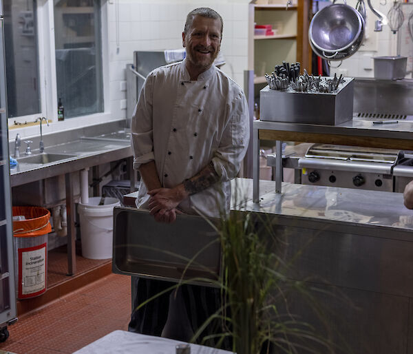 A chef in whites standing behind a decorated table set in front of a commercial kitchen, smiling to camera