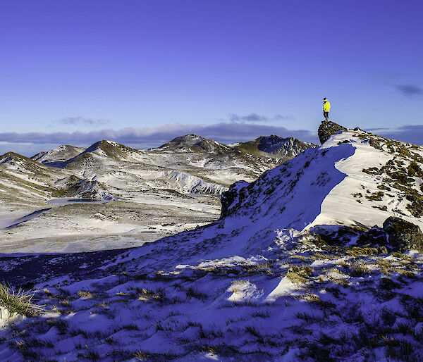 A spectacular snow dusted view across the mountains.  A man stands at the highest point looking out across the amazing landscape.