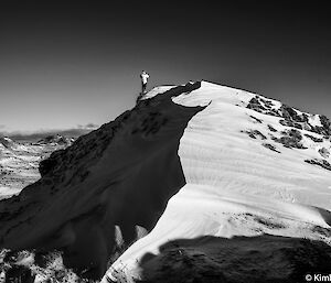 A black & white image looking across a spectacular snow dusted mountain ridge.  A man standing at the highest point looking out across the amazing landscape