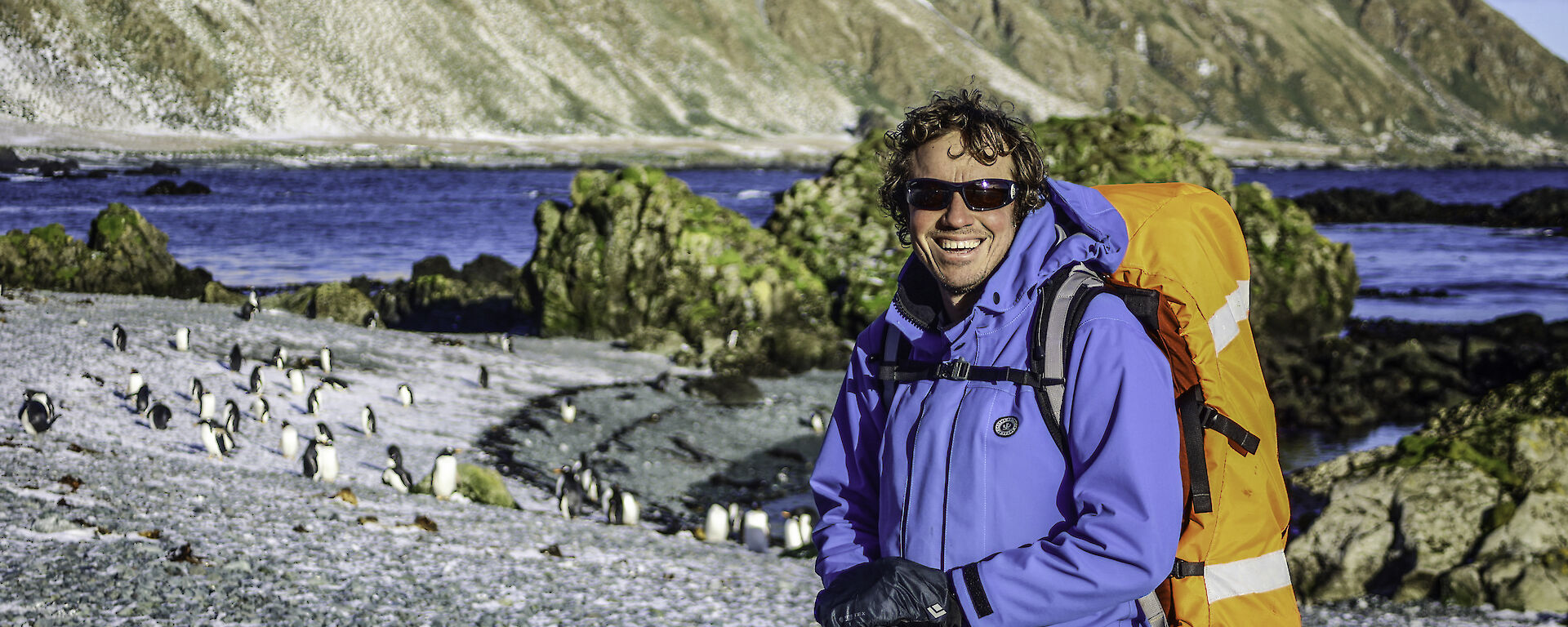 A man with a backpack and walking gear smiles to camera.  Behind him on the shore are a group of penguins and snow dusted mountains.