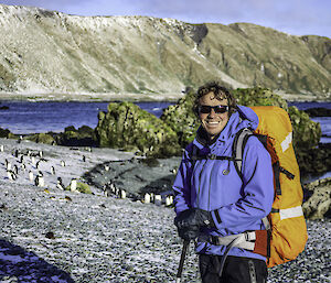 A man with a backpack and walking gear smiles to camera.  Behind him on the shore are a group of penguins and snow dusted mountains.