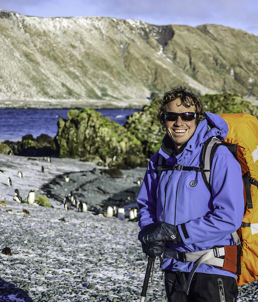 A man with a backpack and walking gear smiles to camera.  Behind him on the shore are a group of penguins and snow dusted mountains.
