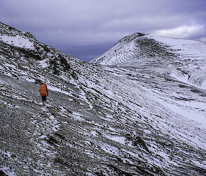 A snowy slope heading towards a peak.  A lone walker can be seen walking away from camera.