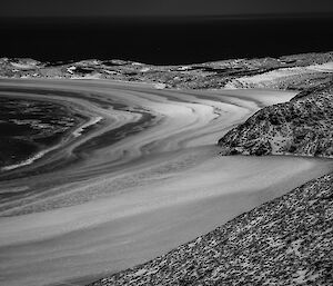 A black and white image of a lake shore with snow dusted hills and frozen wave patterns on the sand