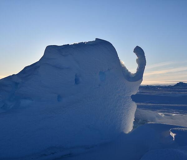 A small ice berg in the shape of an elephants head and trunk with the sun behind it.