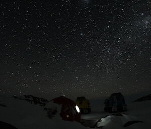 A night photo of the clear starry night with a bright light shining through the round window of the hut.