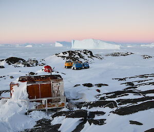 Macey hut in the foreground with the red apple shaped hut along with the blue and yellow Hägglunds near by and then large ice bergs of in the distance.