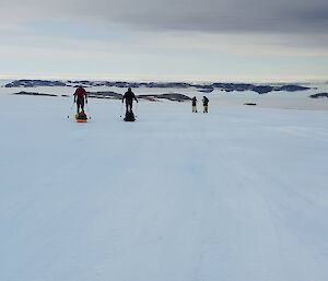 Four expeditioners walking towards the horizon in the snow, each pulling a small sled.