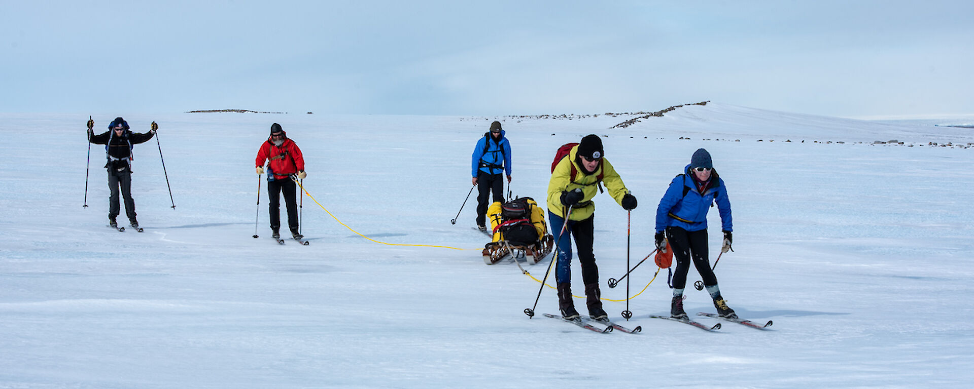5 people on skis on a flat snowy landscape.  Two are attached by rope to a sled.