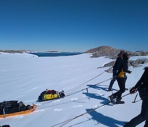 Three expeditioners walk past camera in the snow.  Each of them are pulling a small sled attached to them by rope.
