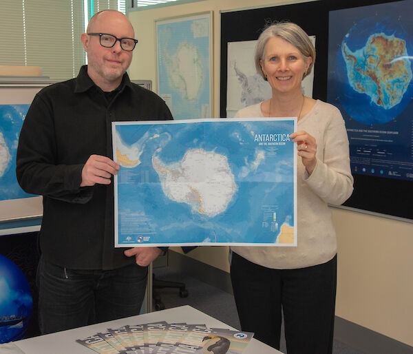A man and a woman hold up a map of Antarctica between them and smile to camera