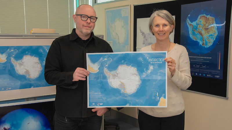 A man and a woman hold up a map of Antarctica between them and smile to camera