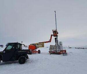 A tall antenna on top of some technical equipment is being moved in place by a small winch.  A Polaris buggy in the foreground and snow on the ground.