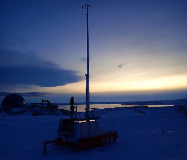 A snowy landscape at dusk.  The silhouette of the weather equipment, particularly a tall antenna can be seen reaching up towards the sky
