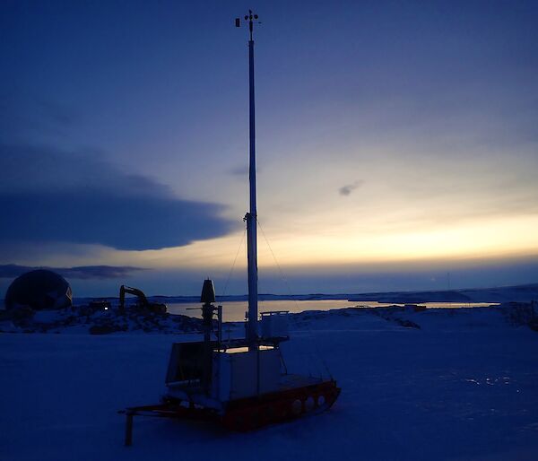 A snowy landscape at dusk. The silhouette of the weather equipment, particularly a tall antenna can be seen reaching up towards the sky