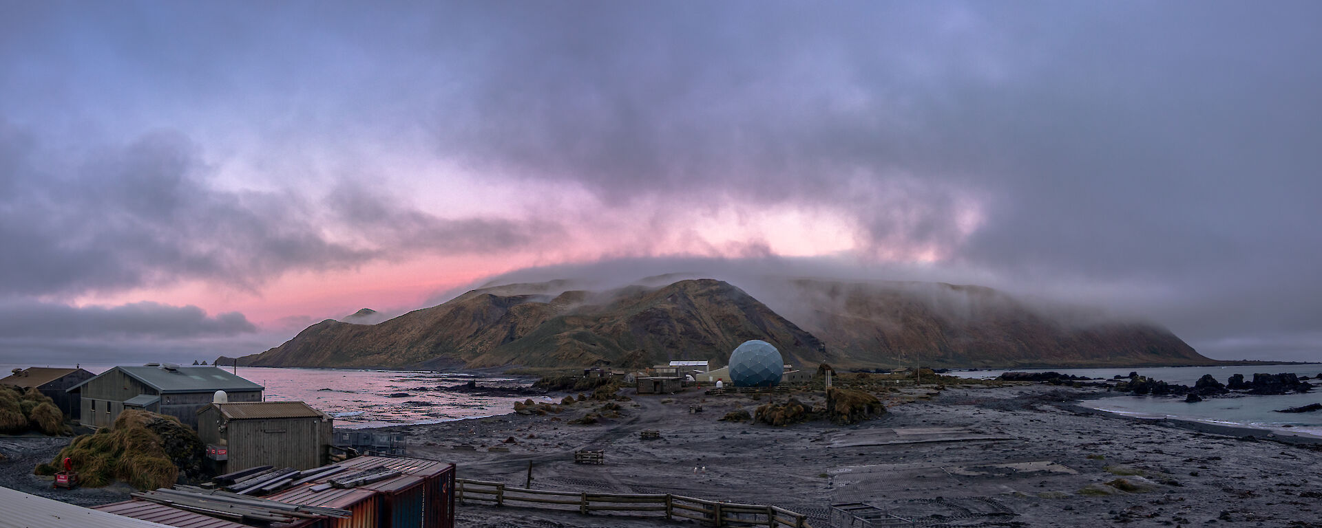 A pick sun sets over a cloudy view of of an isthmus with hills behind