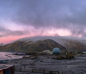 A pick sun sets over a cloudy view of of an isthmus with hills behind