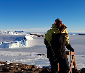 Man in Antarctic clothing standing on a rocky outcrop with ice cliffs in the background.