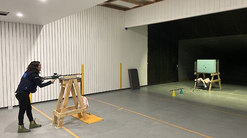 A woman standing behind a dart gun taking aim at a board in the distance.