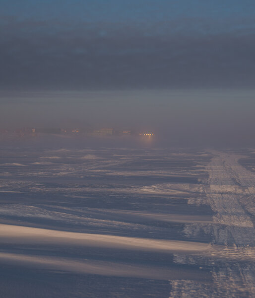 A view across a snowy landscape toward the lights of some distant buildings, just visible through the fog from the sea ice
