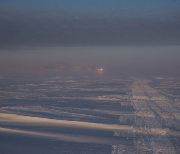 A view across a snowy landscape toward the lights of some distant buildings, just visible through the fog from the sea ice