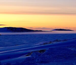 A line of fog across with mountain tops peeking out from above.  The snow below is blue in colour and a yellow sunset lights the sky.