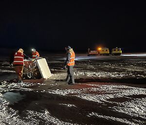 A large crate on the ice with three people around it.  the lights of a Hagglund in the background