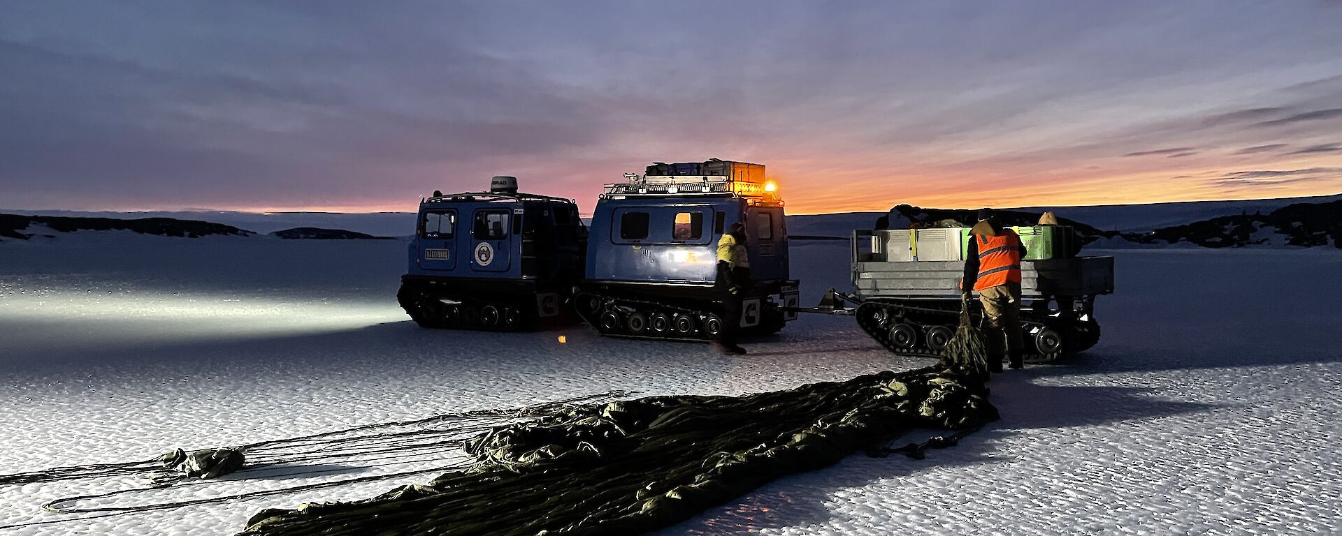 A dusky shot showing a paracute neatly laid on the ice being pulled toward a trailor sat behind a Hagglund