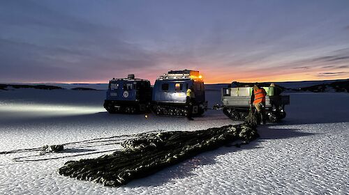 A dusky shot showing a paracute neatly laid on the ice being pulled toward a trailor sat behind a Hagglund