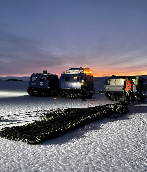 A dusky shot showing a paracute neatly laid on the ice being pulled toward a trailor sat behind a Hagglund