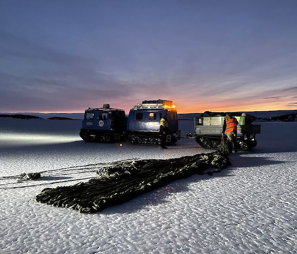 A dusky shot showing a paracute neatly laid on the ice being pulled toward a trailor sat behind a Hagglund