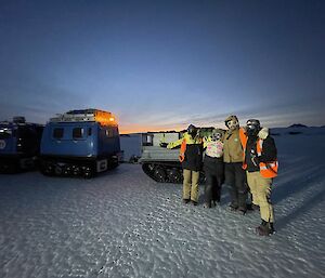 Four expeditioners, warmly wrapped, standing on the ice in front of the loaded Hagglunds smiling to camera