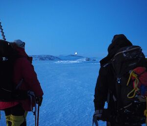 Two people walking away from camera over the sea ice towards the low sun on the horizon.  Both are wearing backpacks and one has a sea ice hole drill sticking out the top.