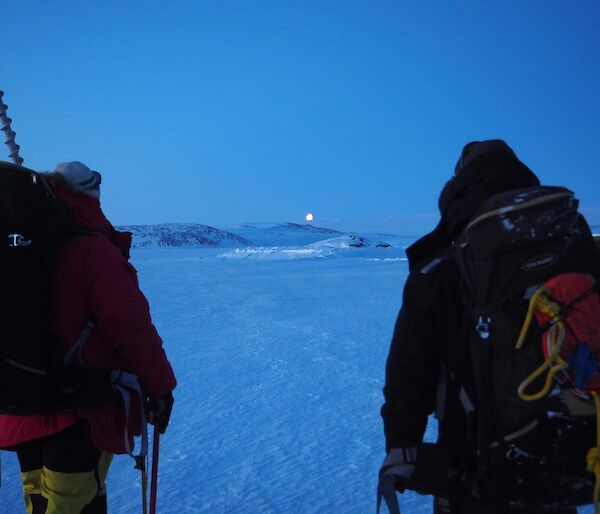Two people walking away from camera over the sea ice towards the low sun on the horizon. Both are wearing backpacks and one has a sea ice hole drill sticking out the top.